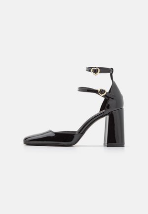 Chaussure à talon haut en cuir verni noir avec deux brides de cheville et boucles dorées en forme de cœur, talon bloc et bout pointu fermé.
