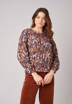 Femme aux longs cheveux bruns portant un chemisier à manches longues marron et noir à motifs et un pantalon couleur rouille, debout devant un fond uni.