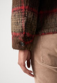 Bruin geruit shirt met rode accenten, gemaakt van een textuur met een wolmix en voorzien van een knoopmanchet. Hand zichtbaar tegen een lichtbeige corduroy achtergrond.