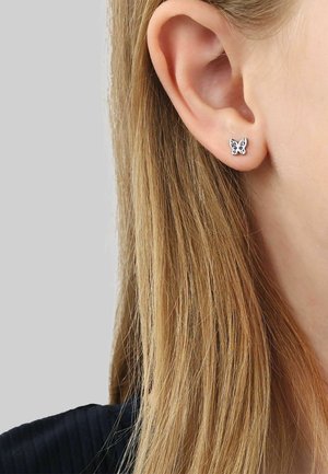 Nahaufnahme eines Ohrs einer Person mit langen blonden Haaren, die einen kleinen silbernen Schmetterlingsstecker trägt.