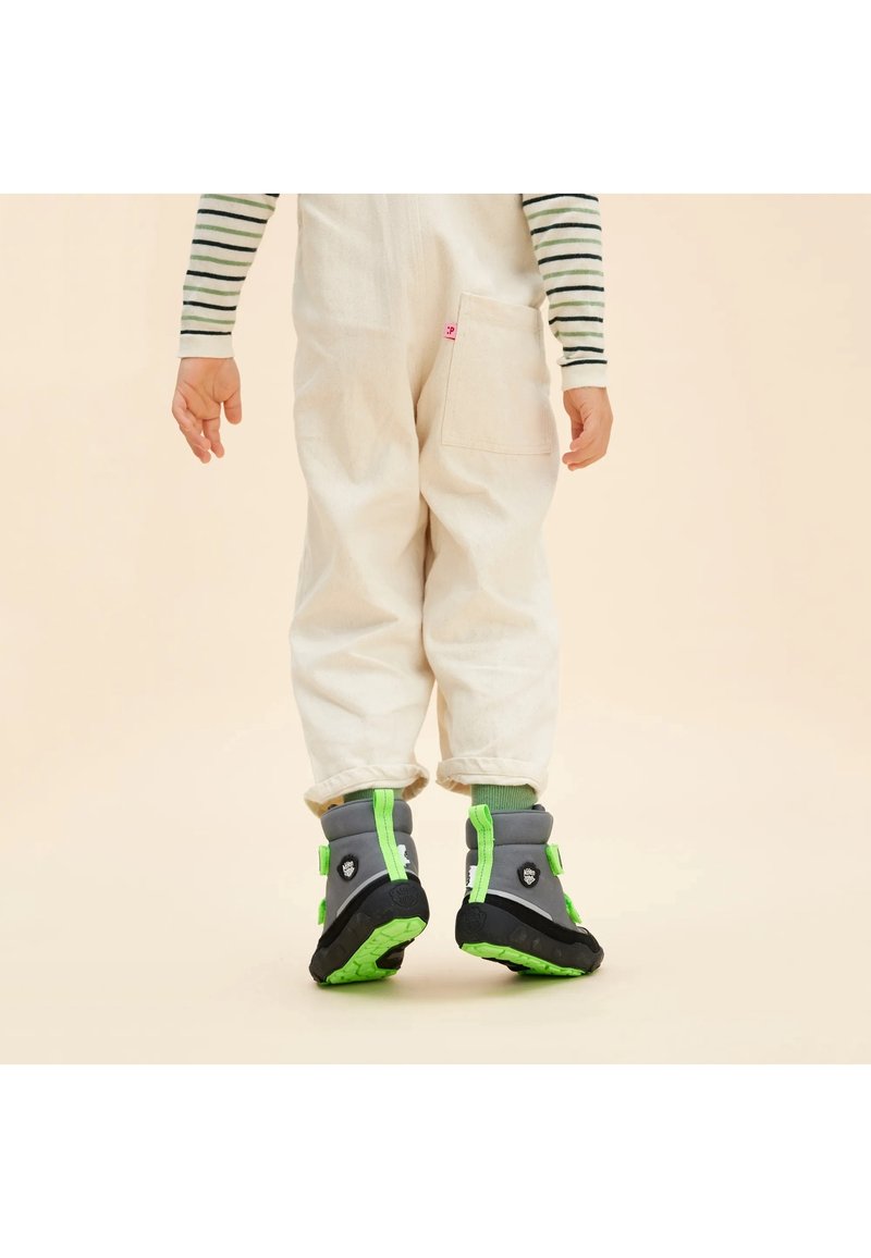 Enfant portant un pantalon blanc et des bottes vertes et grises, debout de dos à la caméra sur fond beige.
