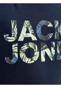 Κοντινό πλάνο ναυτικού υφάσματος με τα γράμματα "JACK JONES" γεμάτα με πράσινα και μπλε τροπικά φυλλώματα.