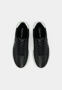 Ζευγάρι μαύρα αθλητικά παπούτσια Calvin Klein με λευκές σόλες και ασημί λεπτομέρειες, απεικονιζόμενα από πάνω σε λευκό φόντο.