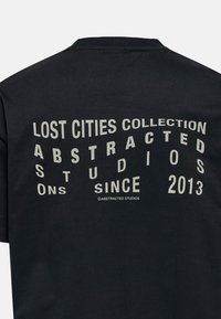 Zwarte t-shirt met gebogen, witte letters op de achterkant. De tekst omvat "Lost Cities Collection" en "Abstracted Studios Sinds 2013."