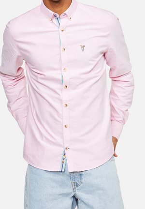 Mężczyzna w jasno-różowej koszuli na guziki z haftem kieliszka koktajlowego i jasno-niebieskich dżinsach, z rękami za plecami.