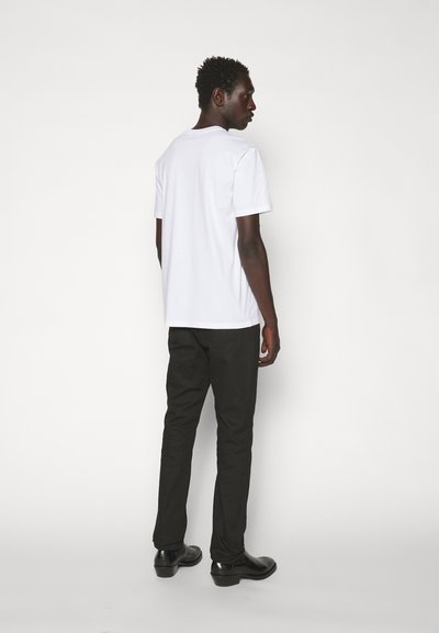 Camiseta blanca de algodón combinada con pantalones negros y zapatos negros, vista desde atrás sobre un fondo blanco liso.