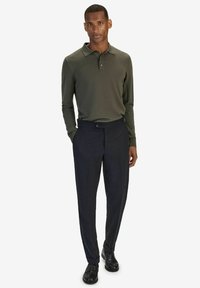 Olijfgroene polo met lange mouwen, gecombineerd met donkere, op maat gemaakte broeken en zwarte schoenen. De stof lijkt glad met een geribbelde textuur.