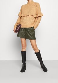 Strickpullover in hellbraun mit Kabelmuster und Rüschensaum. Kombiniert mit dunkelgrünen Ledershorts und schwarzen kniehohen Absatzstiefeln.