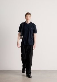 Μπλε ναυτικό μπλουζάκι με στρογγυλή λαιμόκοψη και κοντά μανίκια, σε συνδυασμό με φαρδιά μαύρη παντελόνα. Το μοντέλο κρατά μια μικρή τσάντα χιαστί.