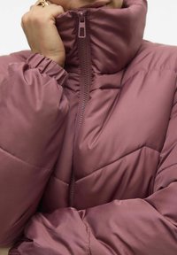 Pufiketas iš švelnaus mauve poliesterio su aukštu apykakle, užtrauktuko uždarymu ir elastingomis rankovėmis. Turi chevrono siuvinėjimo raštą.