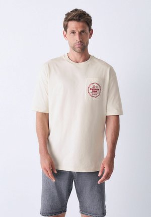 Crème-kleurig t-shirt met korte mouwen, voorzien van een rode zakdesign met tekst. Gemaakt van zachte katoen en heeft een losse pasvorm. Draagt bij denim shorts.