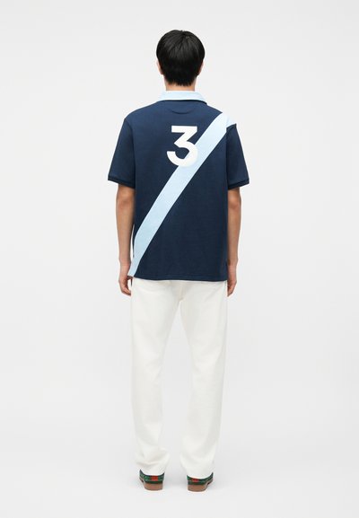 Άνδρας με την πλάτη γυρισμένη φοράει μπλε πουκάμισο πόλο με ανοιχτό μπλε διαγώνια ρίγα και λευκό αριθμό 3, λευκό παντελόνι και σκούρα παπούτσια.