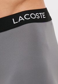 Siv material z gladko teksturo, z črnim pasom in belim logotipom "LACOSTE", ki je jasno prikazan.