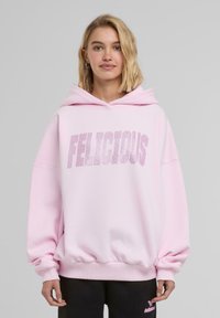 Růžová oversized mikina vyrobená z měkké látky, s velkým grafickým nápisem "FELICIOUS" zdobeným kamínky a klokankovou kapsou.