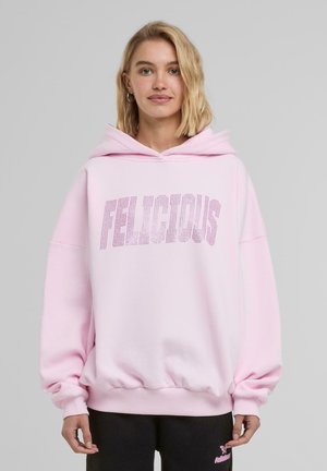 Vaaleanpunainen ylisuuri huppari, joka on valmistettu pehmeästä kankaasta. Siinä on suuri "FELICIOUS" -grafiikka strasseilla sekä kengurupocket.
