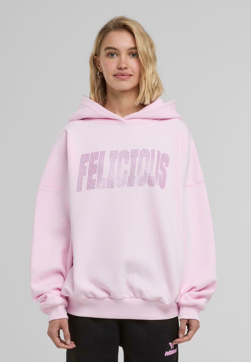 Růžová oversized mikina vyrobená z měkké látky, s velkým grafickým nápisem "FELICIOUS" zdobeným kamínky a klokankovou kapsou.