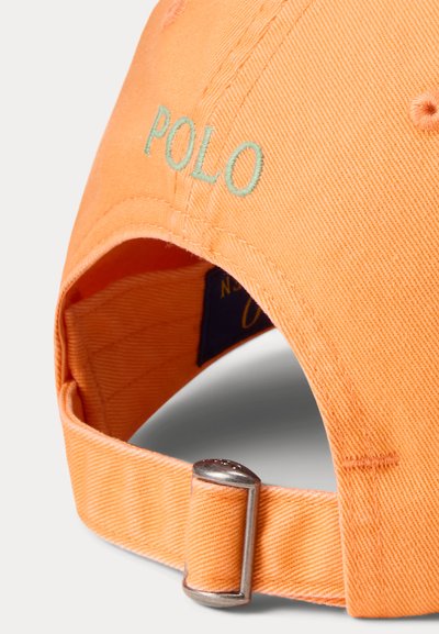 Oranžs beisbola cepure, kas izgatavota no kokvilnas; iezīmēts ar izšūtu "POLO" logotipu gaiši zaļā krāsā. Aizmugurē regulējams metāla jostas sprādze.