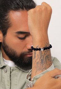 Svart pärlarmband med ett silverdekorerat inslag, bärs på en underarm med synliga tatueringar, mot en ljus bakgrund.
