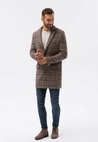 Vesi takki ruskealla ruudullisella kuviolla, johon kuuluu jämäkkä muotoilu, kaksi etutaskua, yhdistettynä sinisiin farkkuihin ja ruskeisiin nahkasaappaisiin nilkkapituisena. Vaalea harmaa villapaita alla.