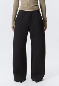 Person som har på seg løstsittende svarte vide bukser og en beige langermet topp, står med hendene hvilende ved sidene, sett bakfra.
