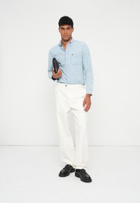 Világoskék farmering zsebbel, fehér nadrág és fekete vastag talpú cipő. A modell egy fekete tárgyat tart a kezében. Egyszerű háttér.