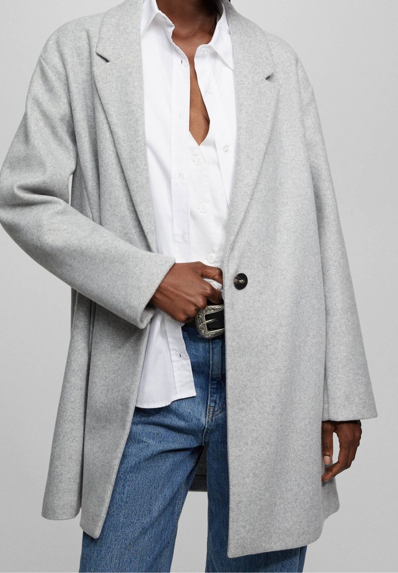Manteau gris en mélange de laine avec une coupe décontractée, de larges revers et un bouton noir unique. Porté sur une chemise blanche boutonnée et un jean bleu.