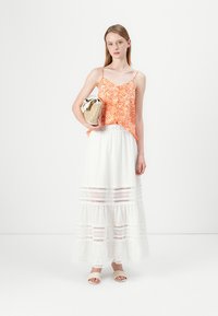 Top cu model portocaliu, fără mâneci, cu bretele subțiri, asortat cu o fustă albă, cu volane și detalii din dantelă. Modelul ține o geantă țesută și poartă sandale tip slide.