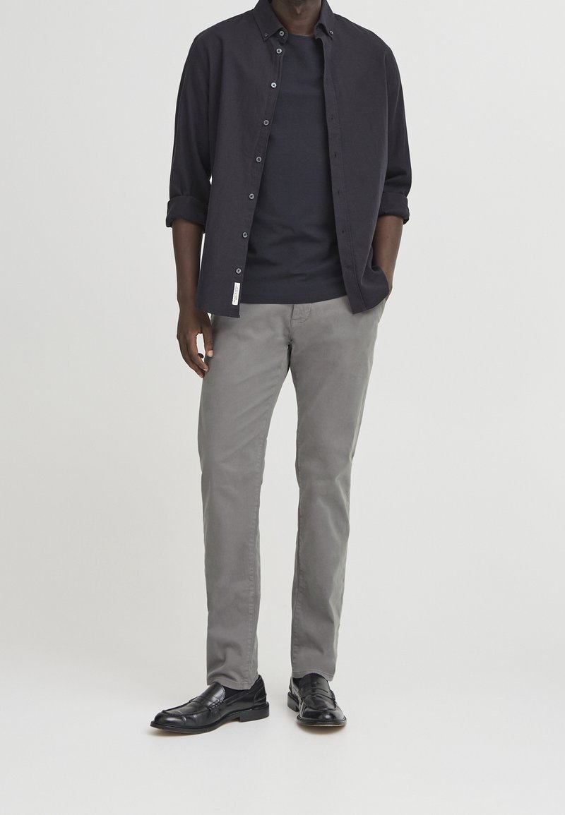 Homme portant une chemise sombre boutonnée avec manches retroussées sur un t-shirt noir, pantalon gris et chaussures habillées noires, debout avec une main dans la poche.