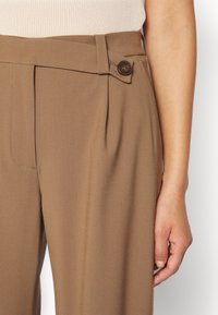 Femme portant un pantalon marron taille haute, ajusté avec une patte boutonnée sur le côté, associé à un haut sans manches beige côtelé.