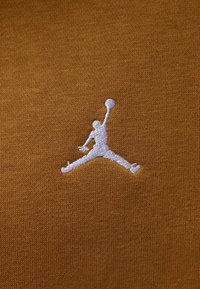 Silueta blanca bordada de un jugador de baloncesto saltando con un balón sobre una tela de punto marrón.