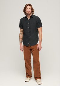 Camicia a maniche corte, grigia scura, con bottoni e tessuto texturizzato, abbinata a pantaloni in velluto a coste marroni. La camicia ha un orlo dritto e un'etichetta sul lato.