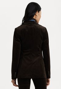 Καφέ παντελόνι τζάκετ με κομψή εφαρμογή, καμπύλες πατιλέτες και λεπτομέρειες με τέσσερα κουμπιά στα μανίκια. Απαλή υφή με κάθετες γραμμές.