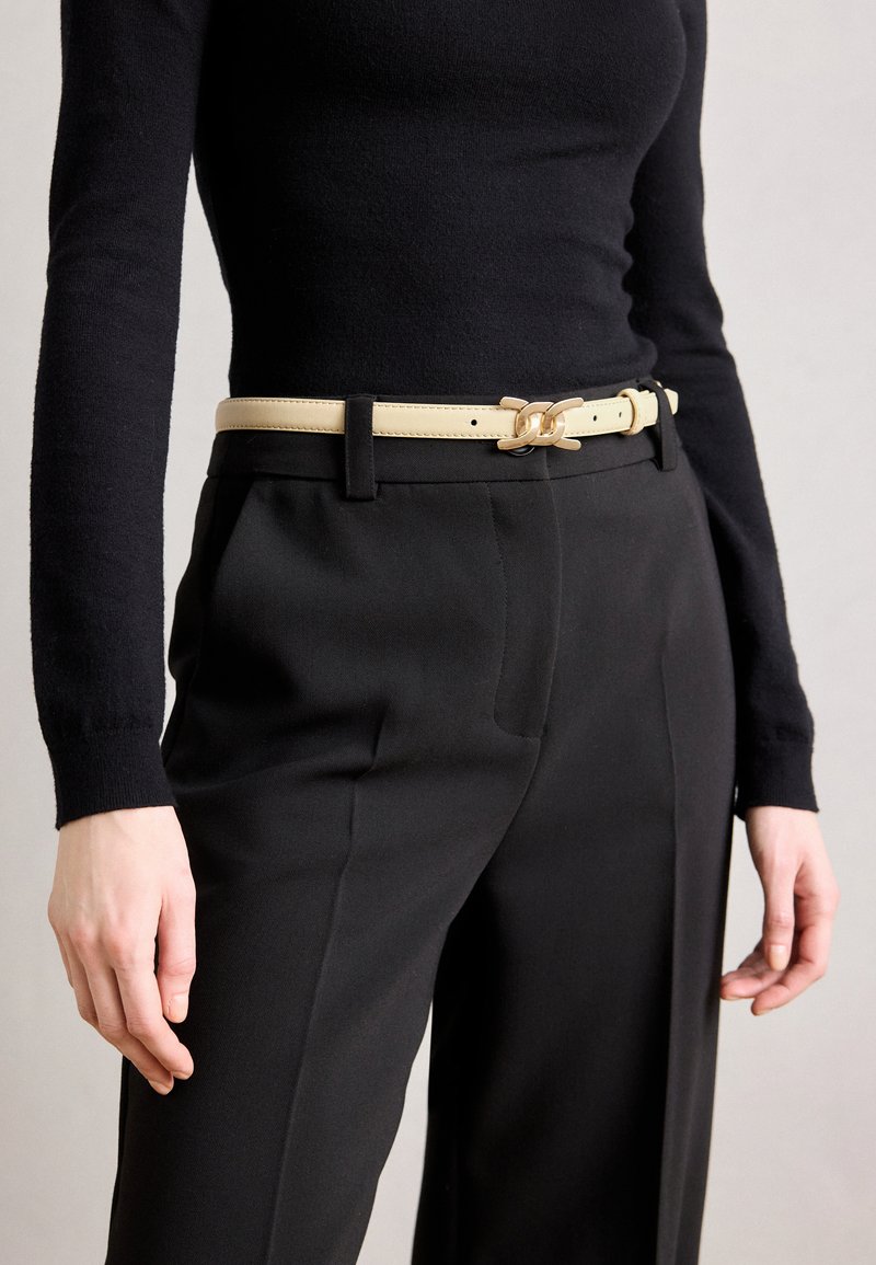 Ceinture en cuir beige avec des éléments en chaîne, portée sur un pantalon noir et un haut ajusté noir. Accent sur le design minimaliste et la texture lisse.