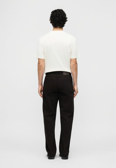 Άνδρας με σγουρά σκούρα μαλλιά στέκεται με την πλάτη γυρισμένη, φορώντας απλό λευκό κοντομάνικο πουκάμισο και φαρδιά μαύρη παντελόνια με μαύρα παπούτσια.