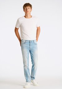 Heledad sinine teksapüksid kulumisega viimistluses, millele on sobitatud lihtne valge T-särk. Mudel seisab rahulikult käed taskus. Valged tossud.