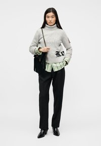 Nainen, joka pukeutuu harmaaseen pooloneuleeseen vihreän raidetun paidan päällä, mustiin housuihin, mustiin nilkkureihin ja kantaa mustaa hapsulaukkua.