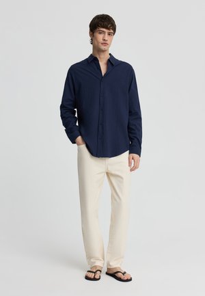 Giovane uomo che indossa una camicia blu scuro con bottoni, pantaloni color crema e infradito nere, in piedi con una mano in tasca su uno sfondo semplice.