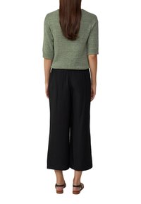 Femme aux longs cheveux bruns portant un pull en tricot vert, un pantalon large noir et des sandales noires, se tenant de dos sur un fond blanc.