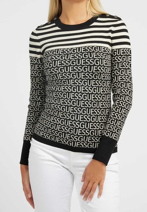 Mujer con un jersey de rayas negras y blancas con el texto repetido "GUESS" y botones plateados en los hombros, combinado con pantalones blancos.