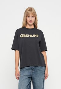 Pelēks pārāk liels T-krekls ar apaļu kakla izgriezumu un īsām piedurknēm, uz kura priekšpuses izcila bežīgu "Gremlins" logotips.