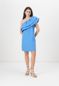 Robe bleue à une épaule avec des volants en couches, longueur au-dessus du genou, fabriquée dans un tissu lisse, assortie à des talons beiges à lanières.