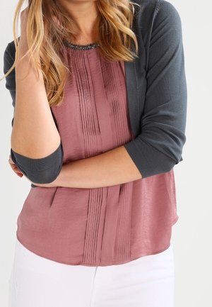 Kvinde med langt blondt hår iført en mauve plisseret bluse, en mørkegrå cardigan og hvide bukser, armene krydset foran livet.