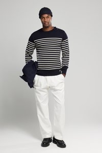 Marineblau- und creme geringelter Pullover mit Knopfdetails an der Schulter, kombiniert mit weißen Hosen und schwarzen Schuhen, während er eine marineblaue Jacke hält.