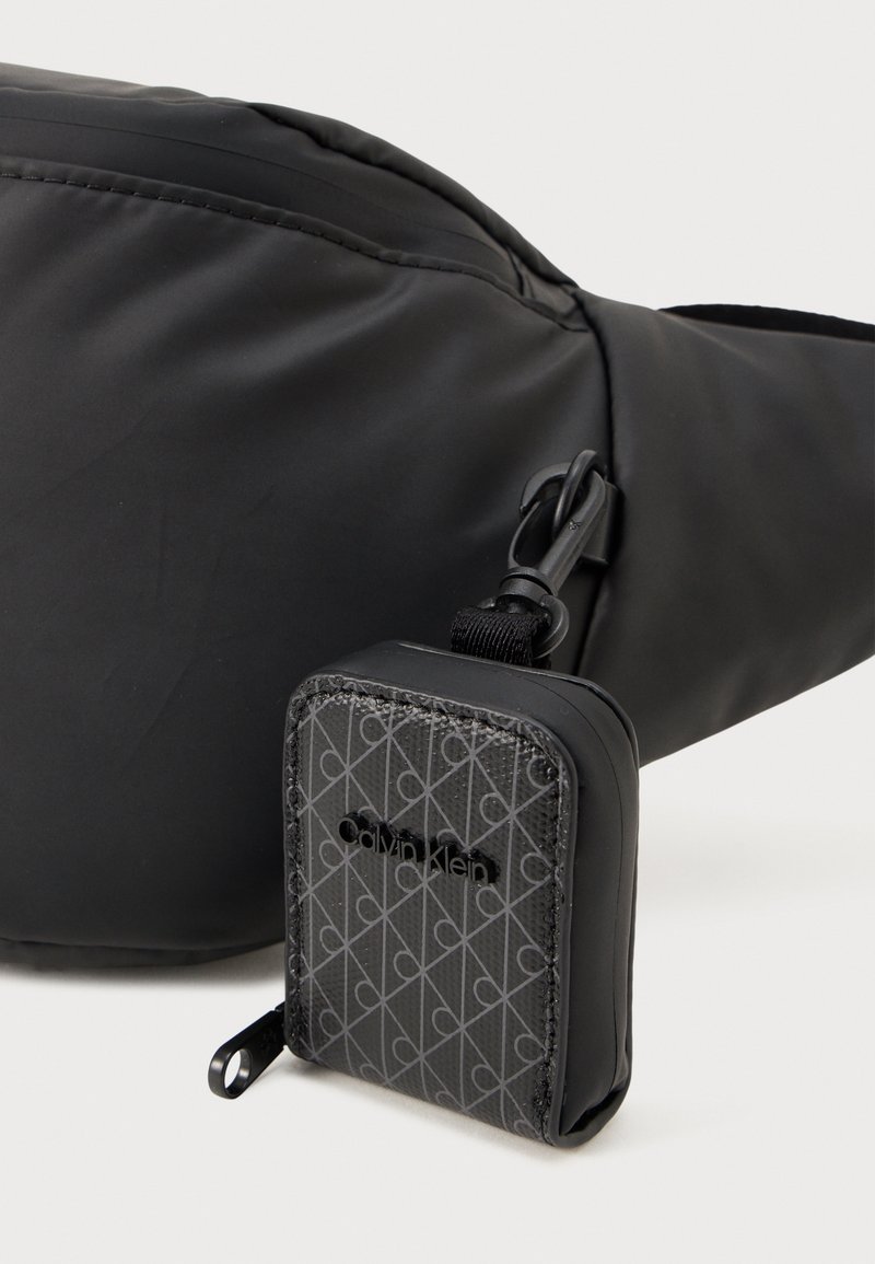 Sac en bandoulière noir en tissu avec une texture lisse, comprenant une petite pochette détachable avec un imprimé géométrique et un logo Calvin Klein.