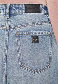 Jean en denim bleu clair à taille haute, avec des coutures orange et une étiquette en cuir noire arborant le logo "ARMANI EXCHANGE" au dos.