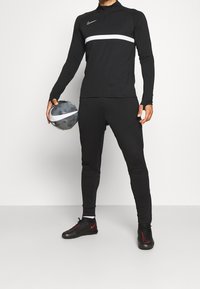 Black athletic outfit featuring a long-sleeve top with a white stripe and fitted pants, holding a soccer ball. Includes black footwear.