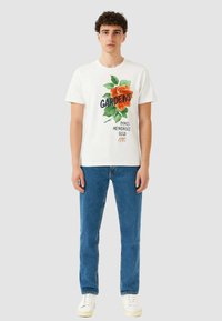 T-shirt bianca in cotone con grafica di rosa arancione e testo "GARDENS". Abbinata a jeans in denim blu e sneaker bianche. Design semplice e casual.