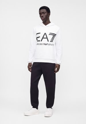 Νέος άνδρας φοράει λευκό φούτερ EA7 Emporio Armani, μαύρο παντελόνι και λευκά αθλητικά, στέκεται μπροστά σε απλό λευκό φόντο.