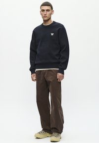 Marineblauer Sweatshirt mit gerippten Bündchen und Saum, versehen mit einem kleinen weißen Logo auf der Brust. Kombiniert mit braunen Kordhosen und beigen Schuhen.
