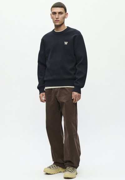 Sudadera azul con puños y dobladillo acanalados, con un pequeño logo blanco en el pecho. Combinada con pantalones de pana marrones y calzado beige.